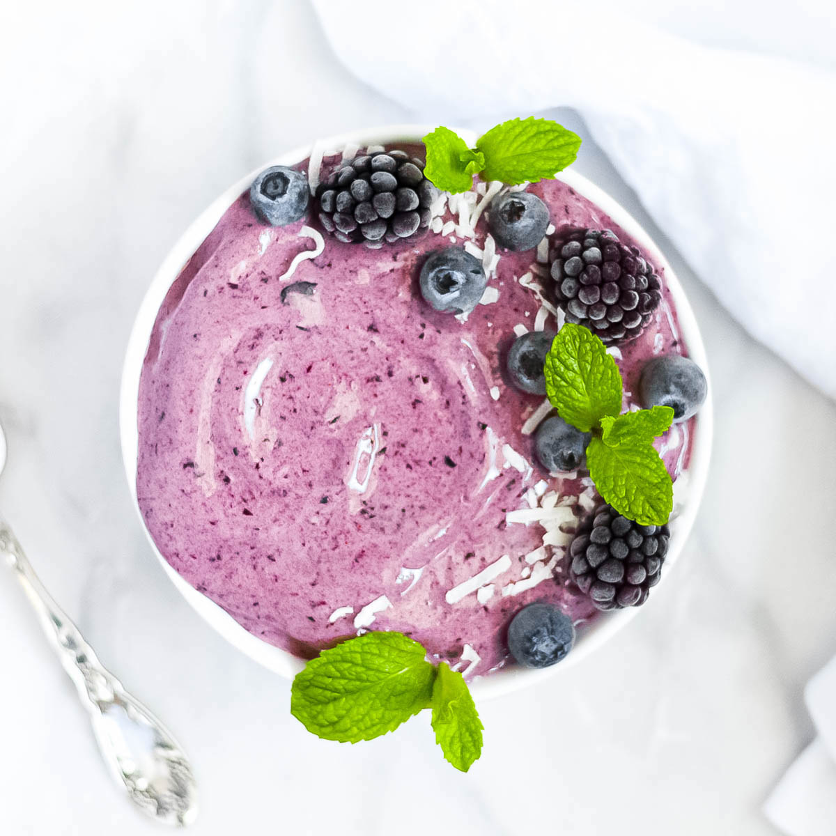 Picture of a purple smoothie in a white bowl, garnished with blackberries, blueberries, coconut flakes, and fresh mint.,