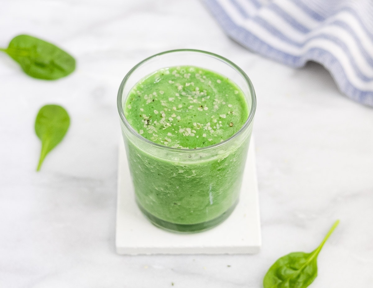 Picture of a green smoothie in a clear glass, sitting on top of a white coaster and surrounded by green spinach leaves and a blue and white dish towel.