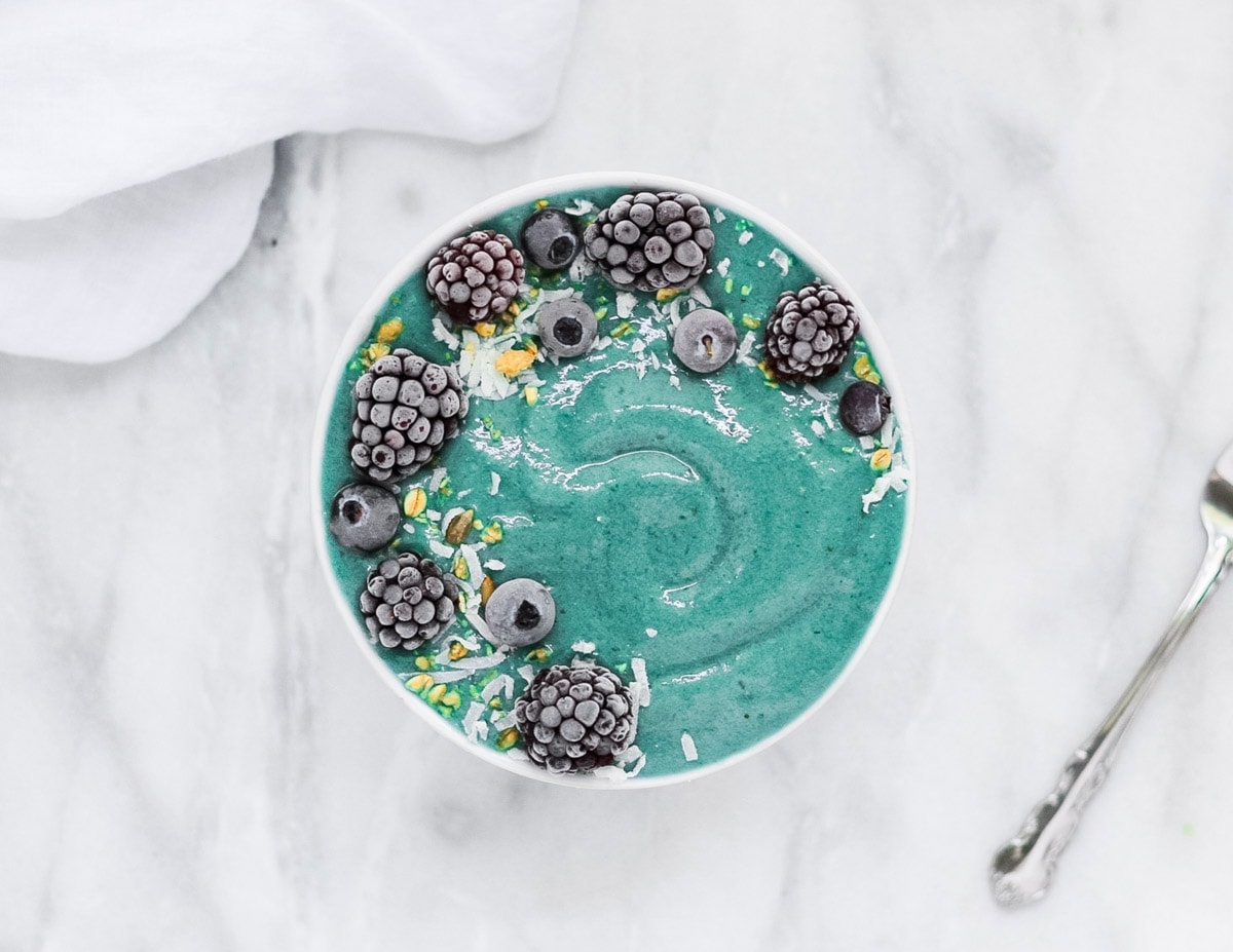 Picture of a blue smoothie in a white bowl topped with granola, coconut flakes, frozen and frosted blueberries and blackberries with a white dish towel and spoon in the background.