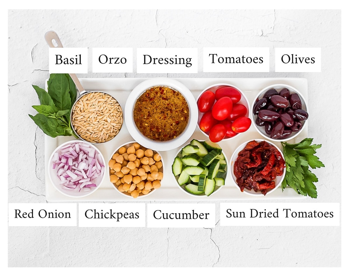 A white platter holding small white bowls of ingredients including: basil, orzo, dressing, tomatoes, olives, red onion, chickpeas, cucumber, sun dried tomato