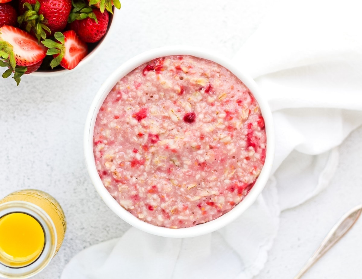 A horizontal picture containing pink raspberry oatmeal. A glass of orange juice, bowl of strawberries, a white cloth napkin, and a spoon are in the background