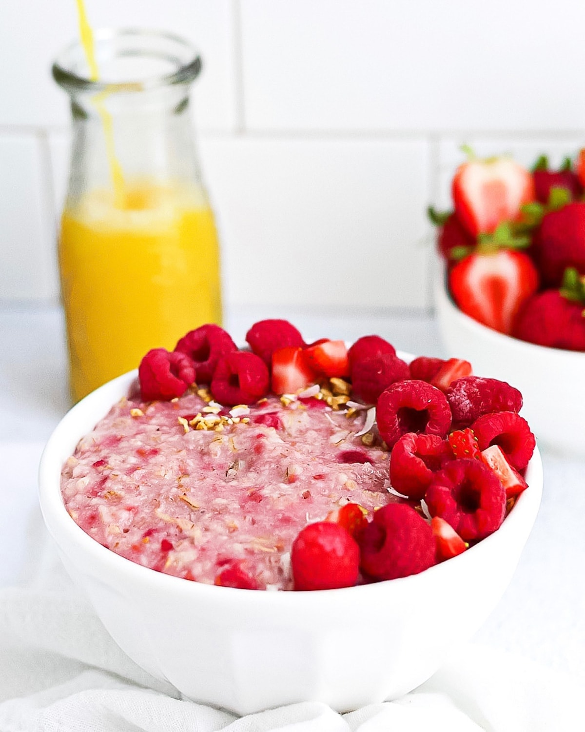 A completed picture of raspberry oatmeal garnish with fruit and granola, orange juice being poured in the background and a bowl of strawberries in the background.