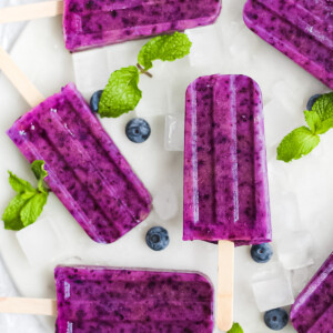 6 purple berry popsicles on a white marble tray, garnished with mint, blueberries, and ice cubes.