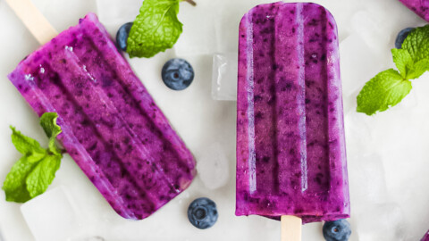 6 purple berry popsicles on a white marble tray, garnished with mint, blueberries, and ice cubes.