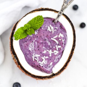 A blueberry smoothie in half of a coconut. Garnished with coconut shreds and fresh mint.