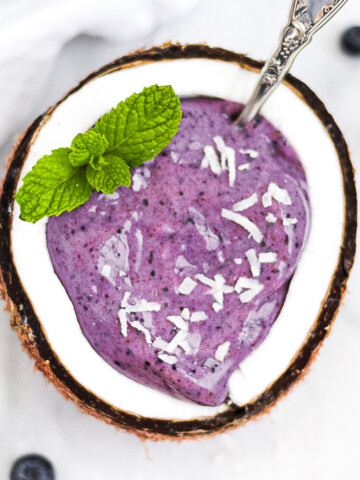 A blueberry smoothie in half of a coconut. Garnished with coconut shreds and fresh mint.