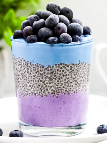 A clear glass holding chia seed pudding parfait, with purple acai yogurt on the bottom, then chia pudding, then blue spirulina yogurt, then a layer of blueberries.