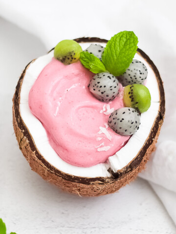 A pink dragon fruit smoothie in a fresh coconut. Garnished with balls of dragon fruit, kiwi, and fresh mint.