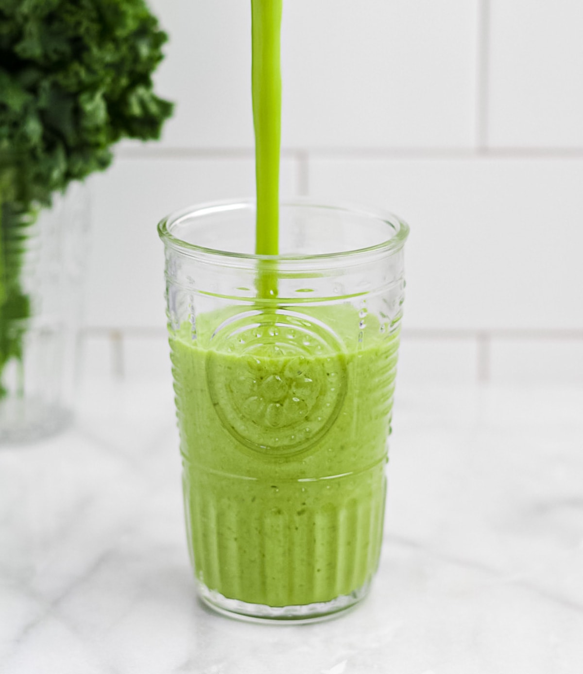 A green smoothie being poured into a clear glass. A bunch of kale is in the background.