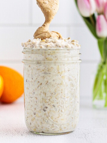 A picture of a clear mason jar with overnight oatmeal inside. A spoon is drizzling peanut butter on top. There are pink tulips and a sliced orange in the background.