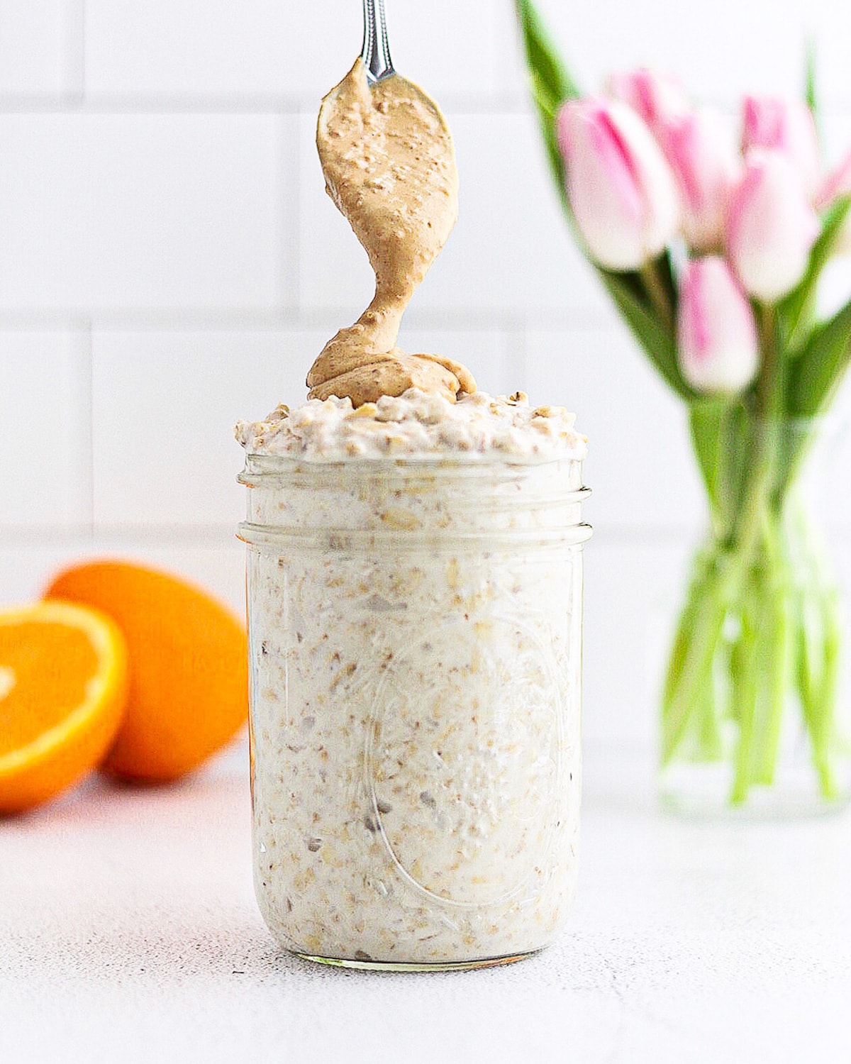 A mason jar with overnight oats inside. There is a silver spoon drizzling peanut butter on top of the oats. There is an orange and a vase of pink tulips in the back.