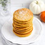 A stack of orange pumpkin pancakes on a stack of white plates. There are pumpkins in the background.