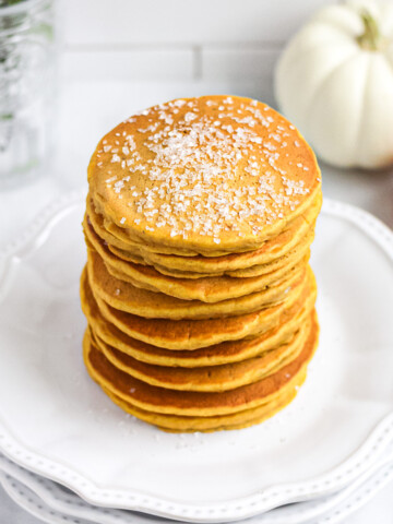 A stack of orange pumpkin pancakes on a stack of white plates. There are pumpkins in the background.