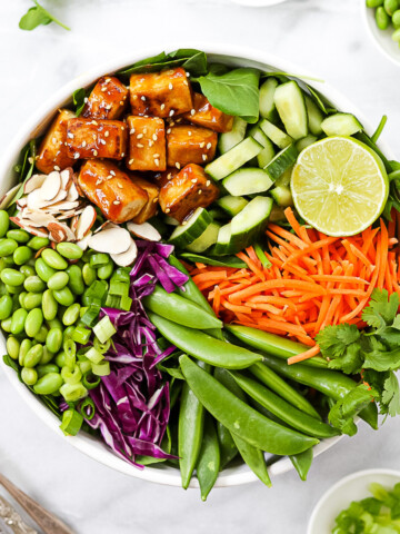 A white bowl holding a large salad, consisting of: sesame tofu, cucumbers, a lime, carrots, snap peas, red cabbage, edamame, and almonds.