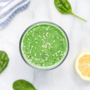 A green smoothie in a clear glass, garnished with white hemp seeds. There is half a lemon, 3 spinach leaves, and a blue and white striped dish cloth in the background.