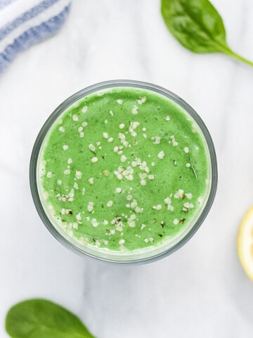 A green smoothie in a clear glass, garnished with white hemp seeds. There is half a lemon, 3 spinach leaves, and a blue and white striped dish cloth in the background.