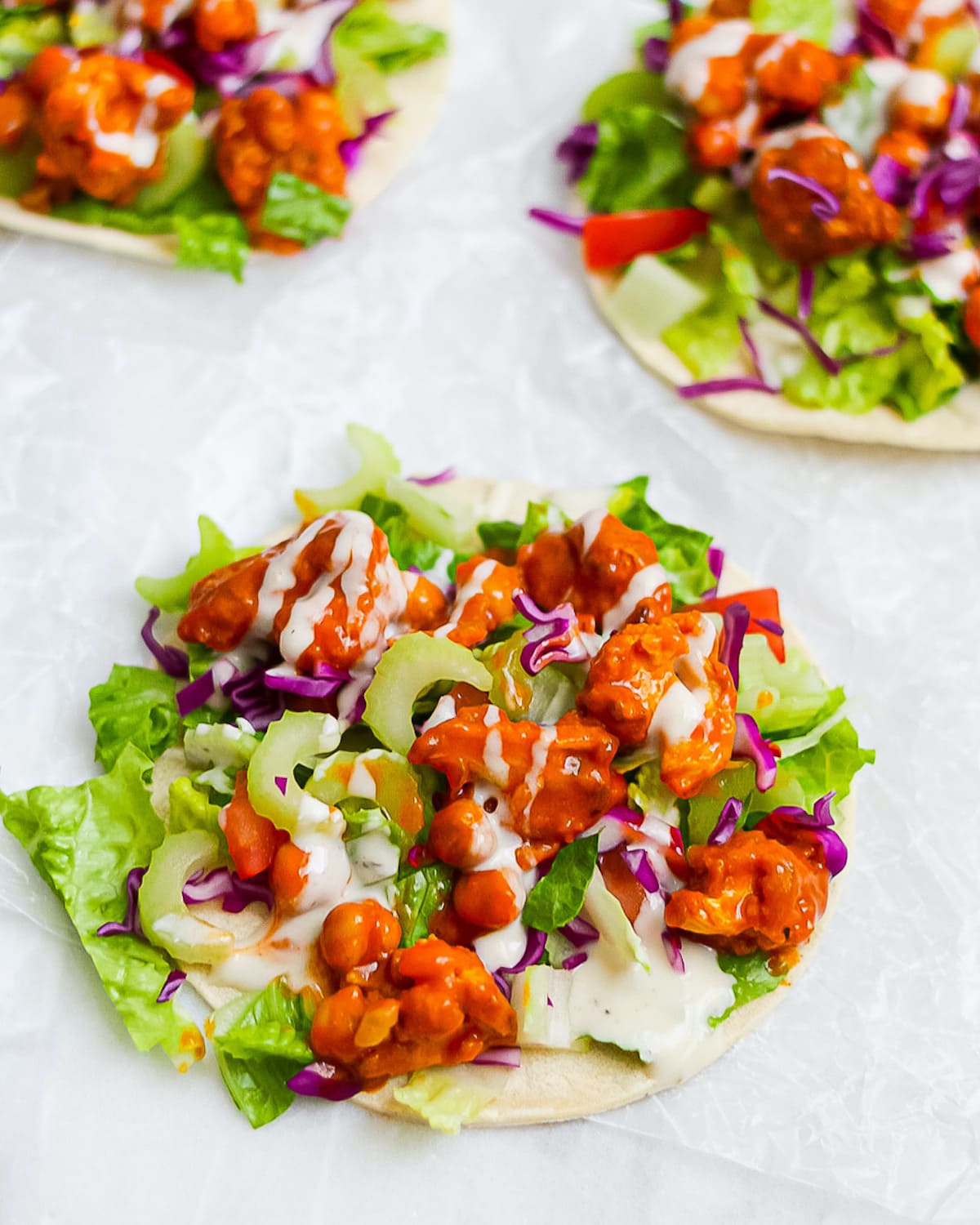 Three tacos with buffalo sauce, cauliflower, chickpeas, lettuce, celery, and tomatoes, with a drizzle of ranch dressing.