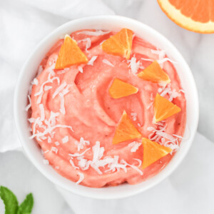 An orange/pink smoothie in a white glass bowl, garnished with coconut flakes and small slices of orange.