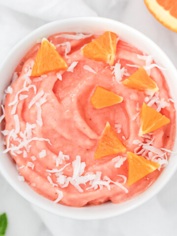 An orange/pink smoothie in a white glass bowl, garnished with coconut flakes and small slices of orange.