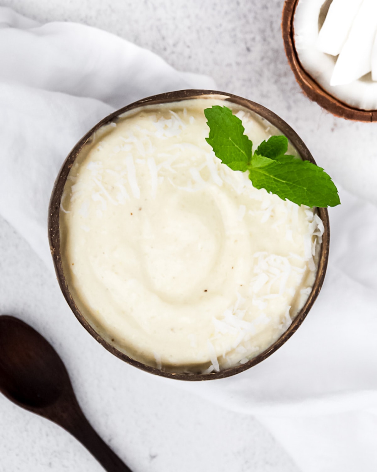 A coconut bowl with a creamy white smoothie inside. It is garnished with fresh mint and white coconut flakes. There is a wooden spoon and fresh coconut in the background.