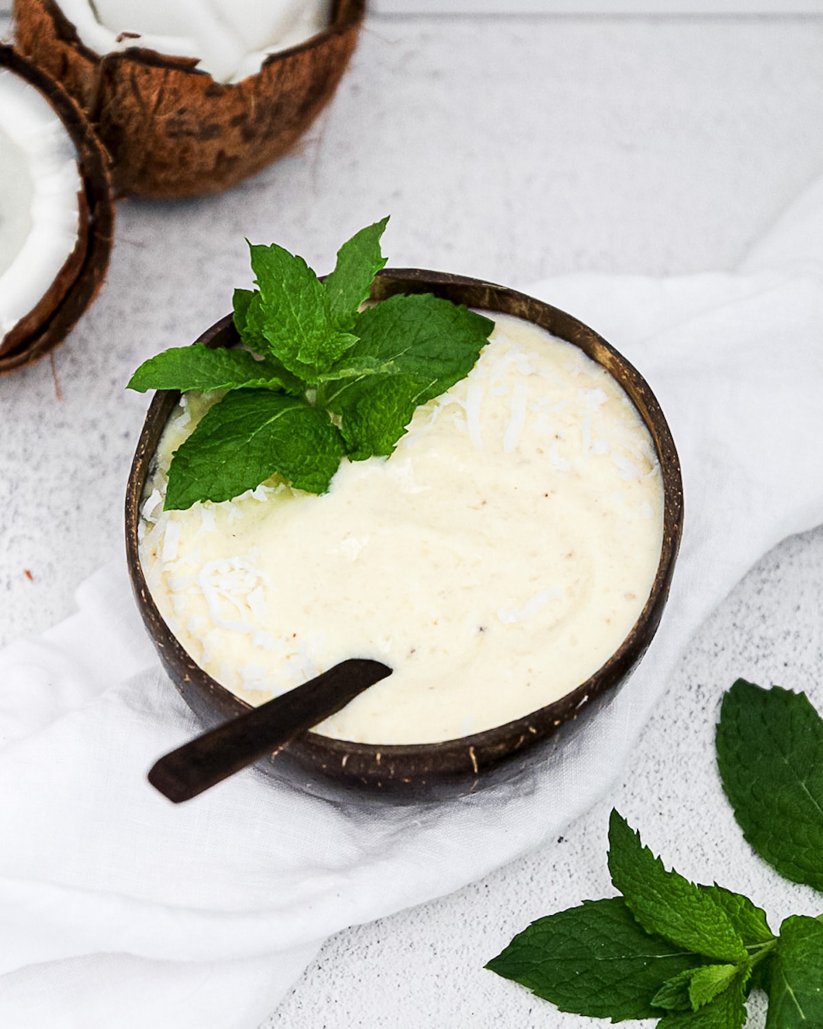 A yellow-white smoothie inside of a brown coconut bowl with a brown wooden spoon in the smoothie. It is garnished with shredded coconut and bright green mint leaves. The bowl is resting on a white linen cloth and there are coconuts and more fresh mint in the corners of the picture.