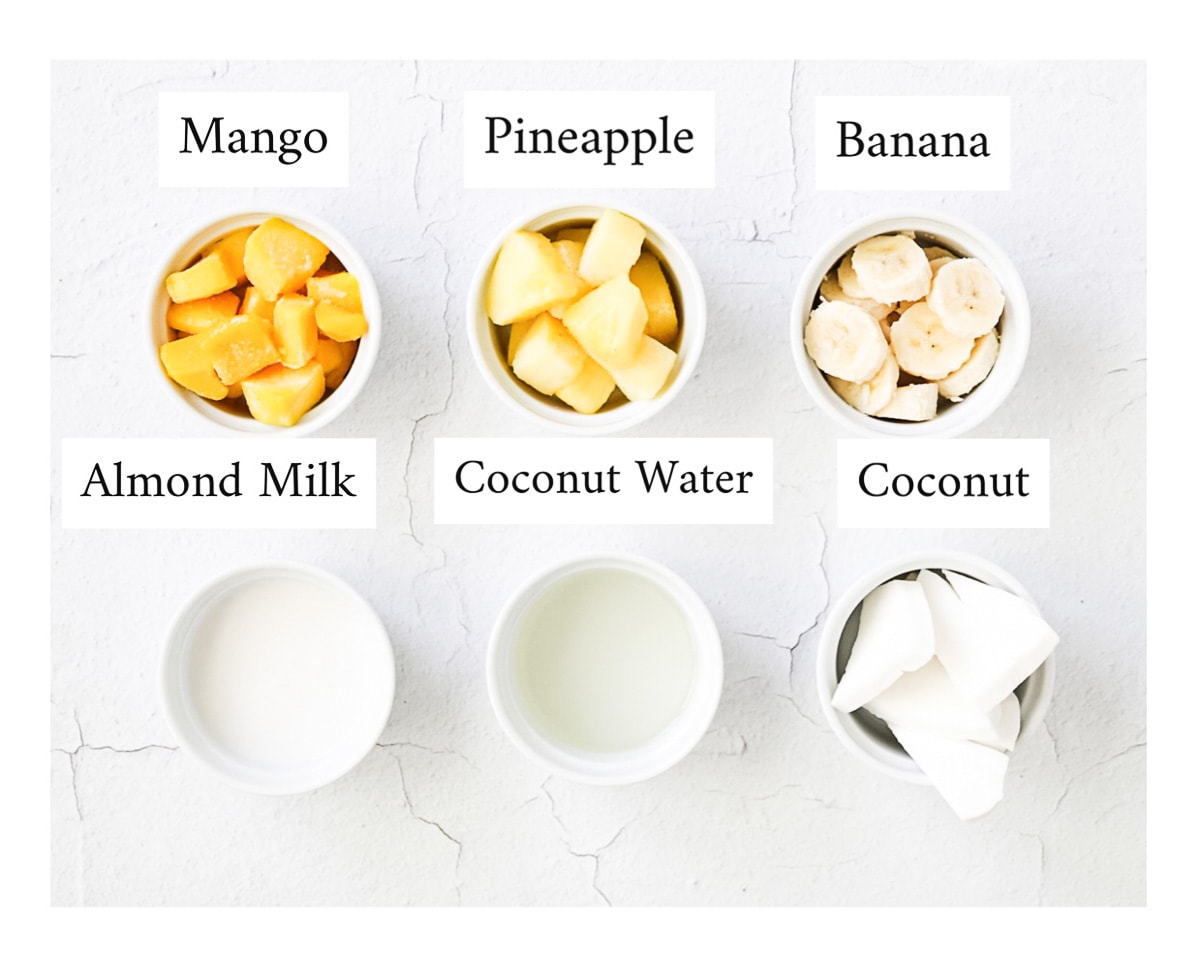 6 small white bowls filled with ingredients and labeled with the ingredient name including: mango, pineapple, banana, almond milk, coconut water, and coconut.