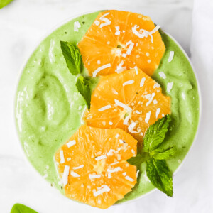 An overhead picture of a bright green spinach smoothie in a white bowl. It is decorated with oranges and green mint leaves, and a sprinkle of coconut flakes.