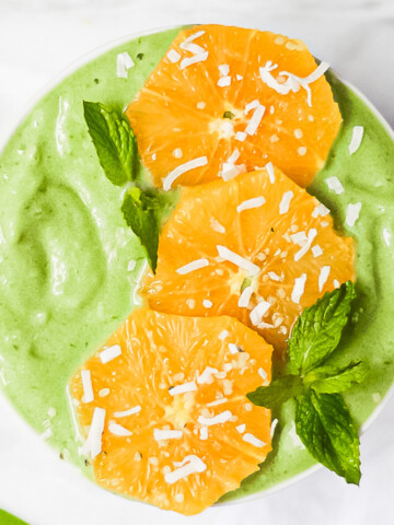 An overhead picture of a bright green spinach smoothie in a white bowl. It is decorated with oranges and green mint leaves, and a sprinkle of coconut flakes.