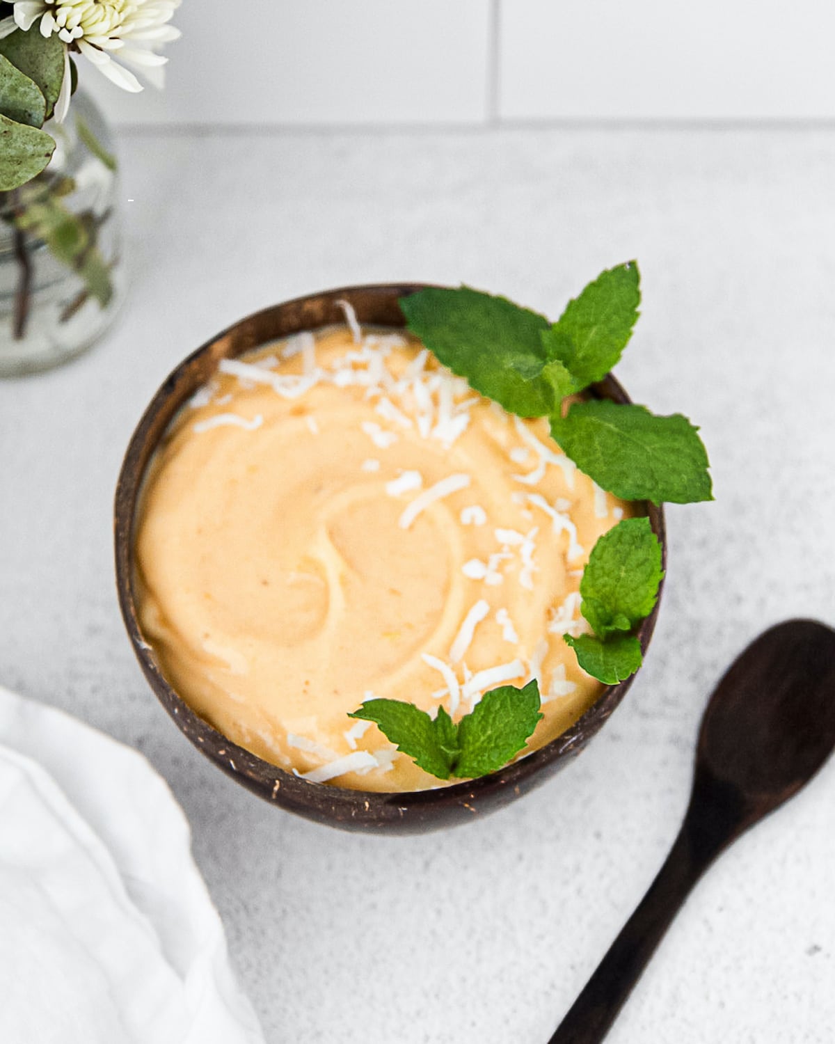 A creamy orange smoothie in a coconut bowl. There is a wooden spoon, vase of flowers, and a white cloth in the background. There are coconut flakes and and mint sprigs in the smoothie bowl.