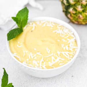 A bright yellow smoothie bowl in a white dish. It is garnished with white coconut flakes and a sprig of light green mint. There is a pineapple, white linen cloth, and more mint in the background.