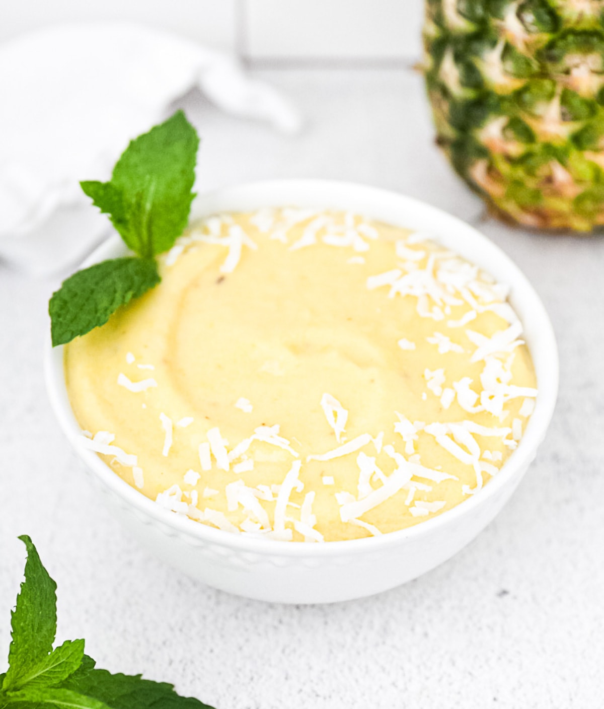 A 45 degree angle picture of a bright yellow smoothie. It is garnished with coconut flakes and mint. There is a white cloth, pineapple, and more mint in the background.