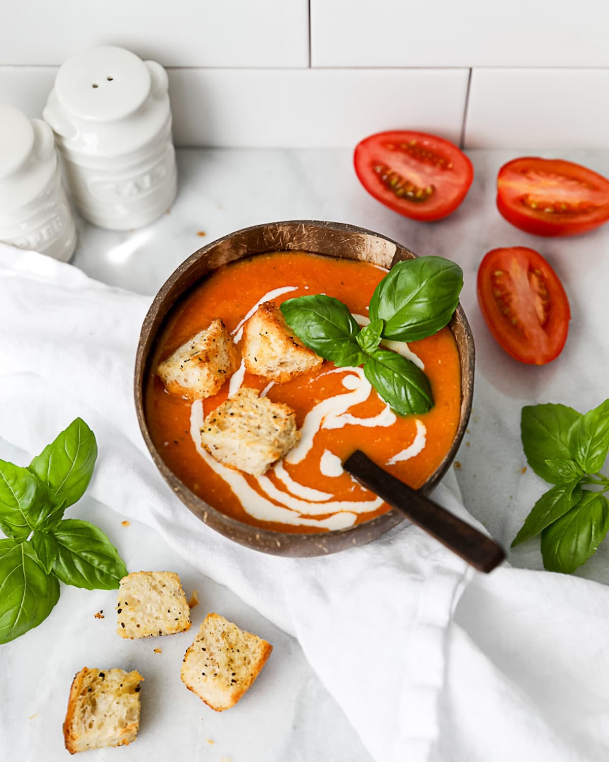 A brown coconut bowl with red soup inside, garnished with vegan cream, three croutons, and fresh basil. There is a wooden spoon inside the soup bowl.