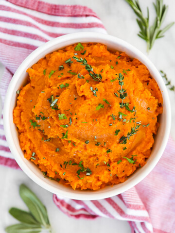 Orange mashed potatoes in a white bowl, garnished with fresh green herbs.