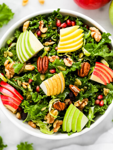 A kale salad in a white serving bowl topped with apples, pears, nuts, pomegranate