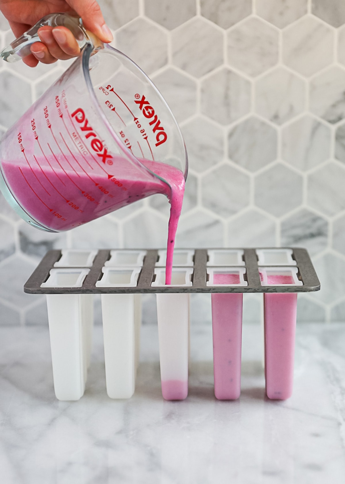 A hand holding a measuring cup that is pouring the unfrozen pink popsicles into popsicle molds.