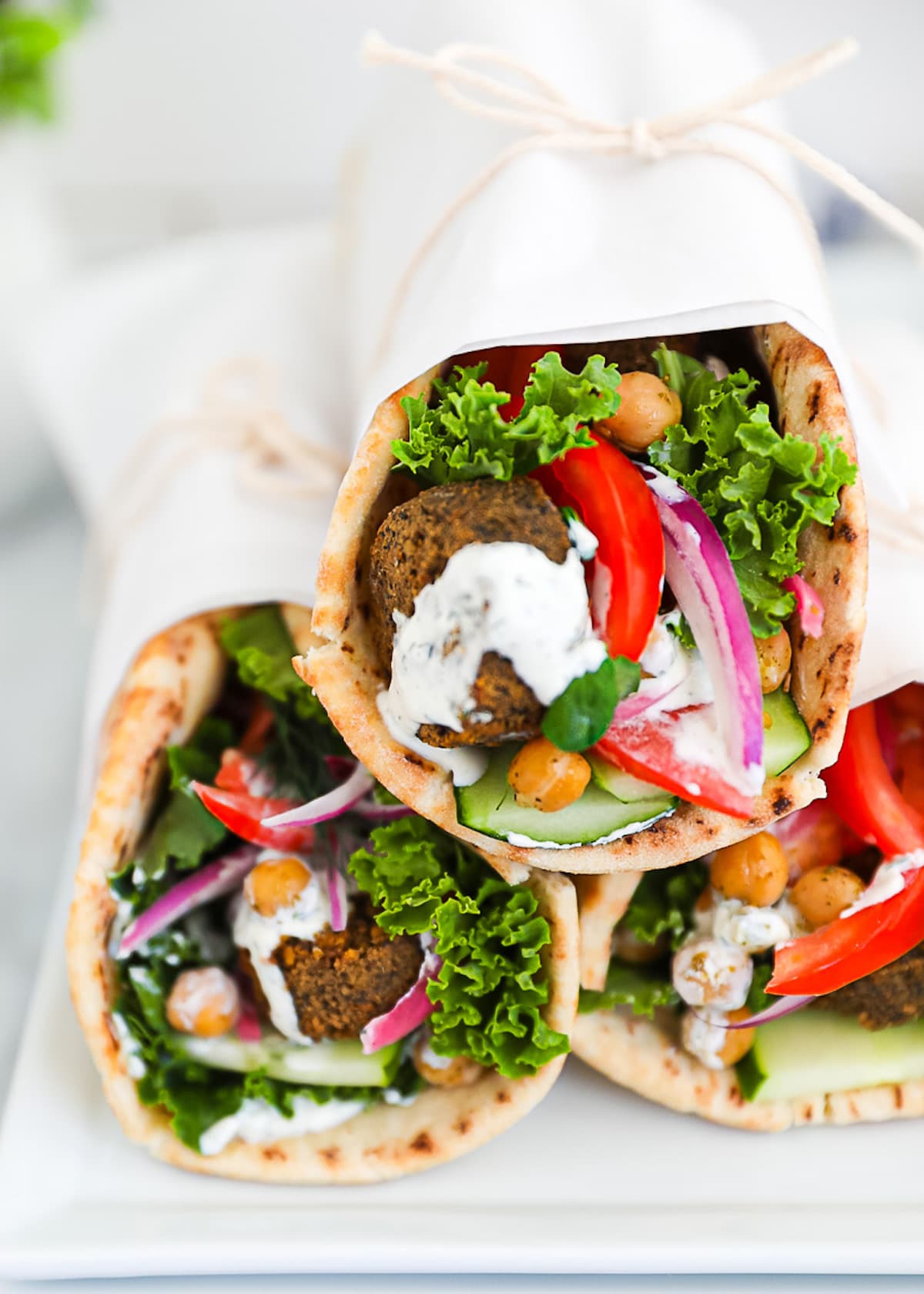 Three vegetarian falafel gyros on a white platter, stuffed with falafel, lettuce, chickpeas, onion, lettuce, tomato, cucumber, and tzatziki sauce.
