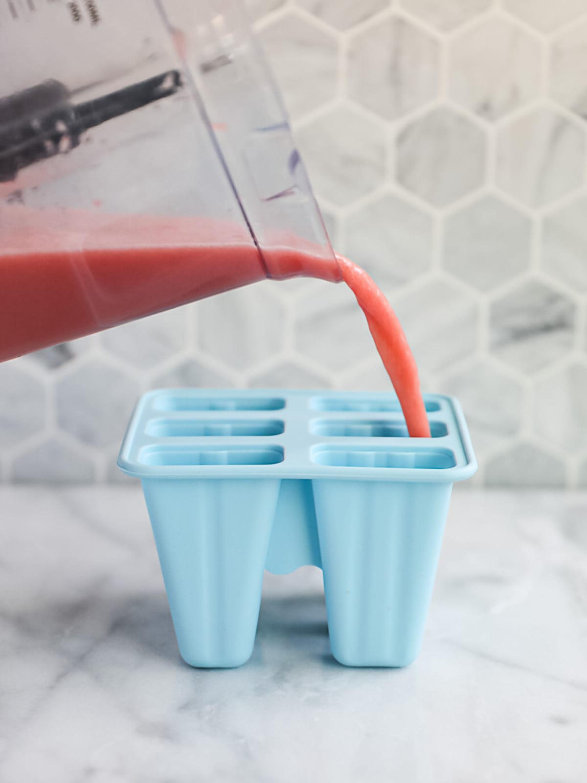 A blender filled with unfrozen watermelon popsicles being poured into blue popsicle molds.