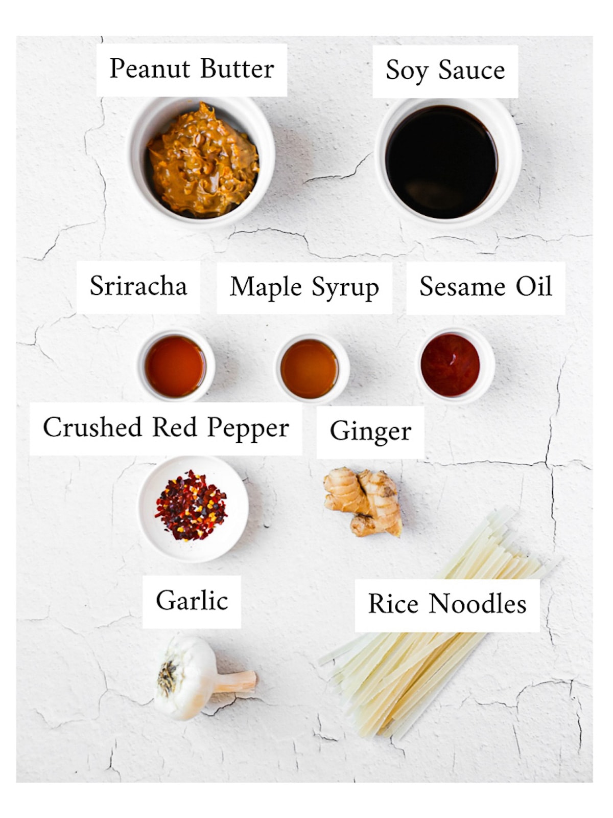Labeled ingredients in small white bowls and on a white background including: peanut butter, soy sauce, sriracha, maple syrup, sesame oil, crushed red pepper, ginger, garlic, and rice noodles.