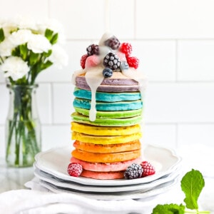 A stack of colorful pancakes with frozen fruit on top and a drizzle of white sugar sauce on top.