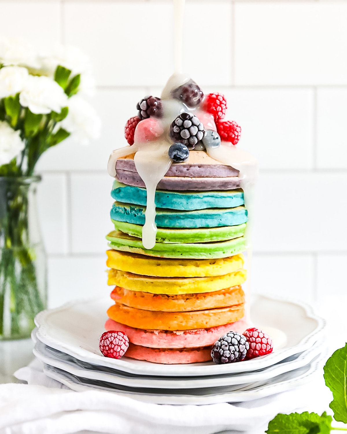 A large stack of rainbow pancakes. There is frozen fruit on top and a drizzle of white cream on top.