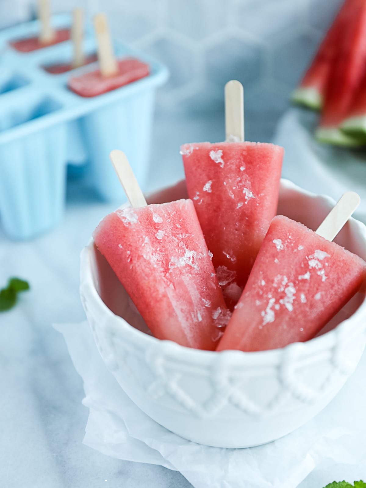 A white bowl of pink watermelon popsicles. There is a popsicle mold and sliced watermelon in the background.