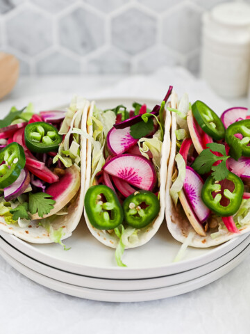 Three vegetable tacos on a stack of white plates filled with fresh vegetables.
