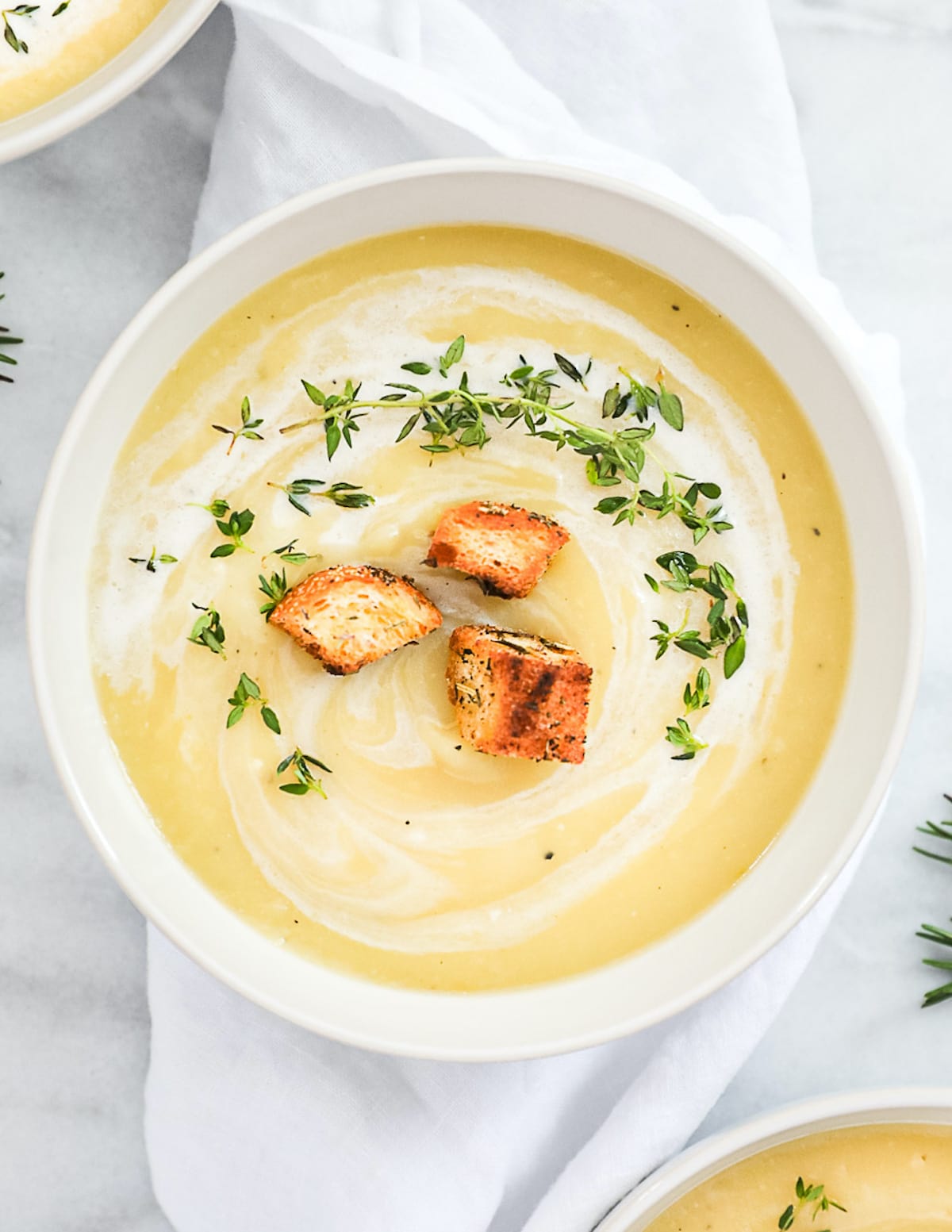 A white bowl filled with creamy yellow soup with a white coconut milk drizzle and fresh herbs and croutons on top