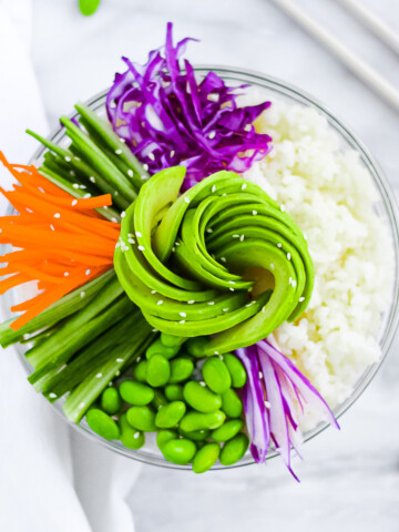 A large bowl filled with a layer of white rice and then shredded red cabbage, sliced cucumbers, sliced carrots, edamame, and a swirled avocado covered in sesame seeds.