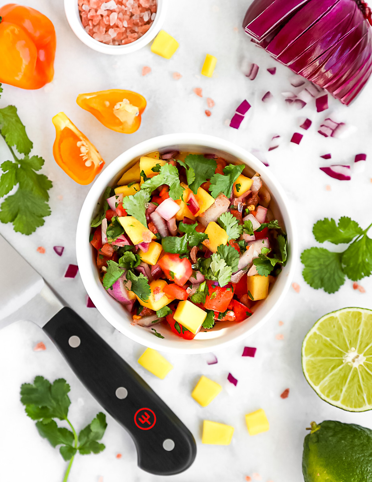 Pineapple mango salsa in a white bowl with the ingredients including: onion, salt, peppers, cilantro, mango, pineapple, and lime scattered around.