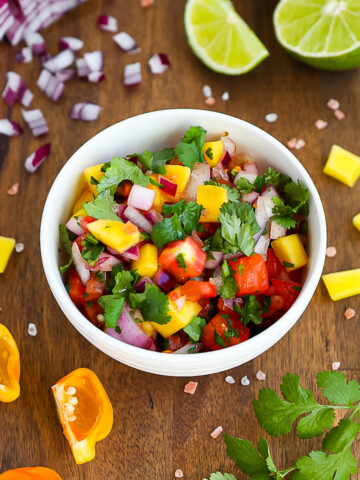 A white bowl sitting on a dark wooden cutting board, the bowl is filled with bright and colorful homemade mango salsa. Ingredients from the salsa are scattered on the cutting board.