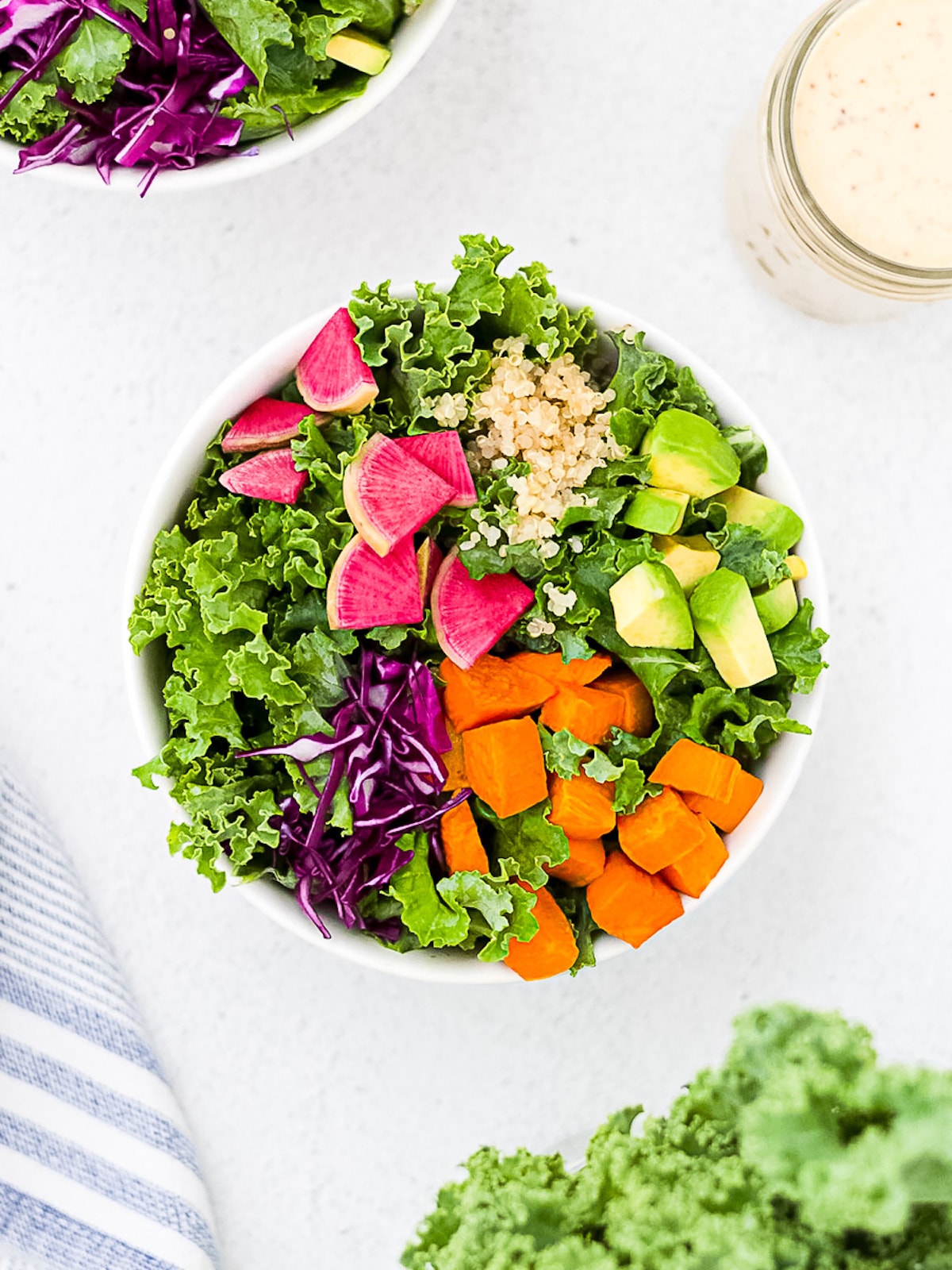 A white bowl filled with prepared kale, pink radishes, sliced avocado, roasted and diced orange sweet potatoes, sliced red cabbage, and cooked quinoa.
