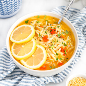 A white bowl filled with lemon orzo soup. Garnished with fresh herbs and three lemon slices.