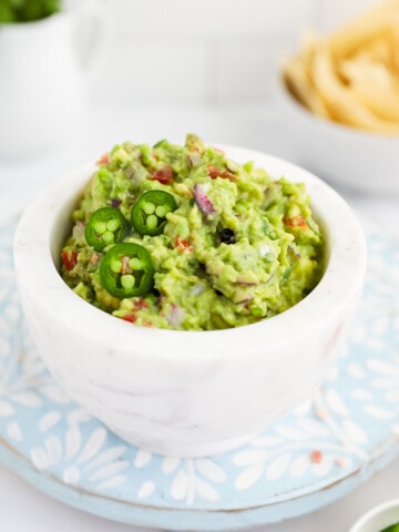 A white marble bowl filled with fresh, bright green guacamole, garnished with three jalapeno slices.