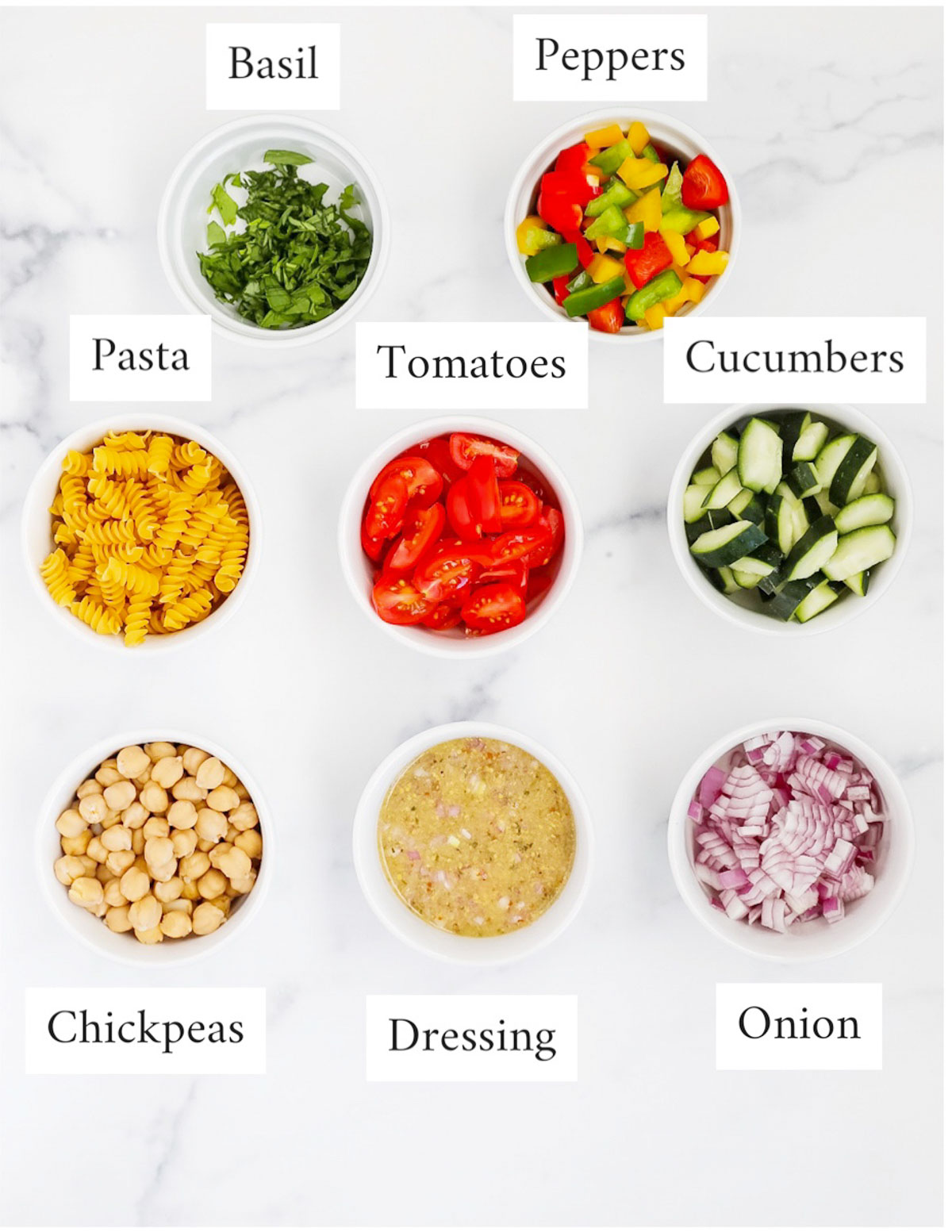 Labeled ingredients in small white bowls including: basil, peppers, pasta, tomatoes, cucumbers, chickpeas, dressing, onion.